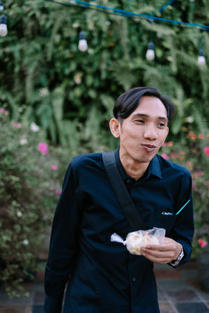 A cheerful man enjoying a snack of street food amidst lush greenery, captured in a candid, lifestyle...