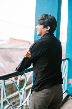 A young man smiles, gazing out over the city from a blue balcony. Contemplative mood.の写真素材