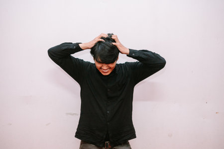 Stressed and frustrated young man pulling his hair. Depicts feelings of despair and being overwhelmed against a plain white background.の写真素材