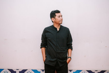 Thoughtful Asian man dressed in black outfit. Casual style with a modern edge, indoor shot near a plain wall.の写真素材