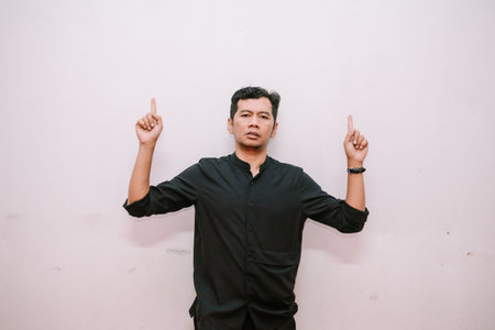Asian man in black shirt pointing upwards to copy space against a plain wall. Great for ads, mockups...