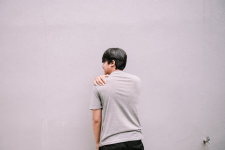 A young man feels discomfort in his shoulder. He touches it, trying to ease the pain. Isolated against a textured grey backgroundの写真素材