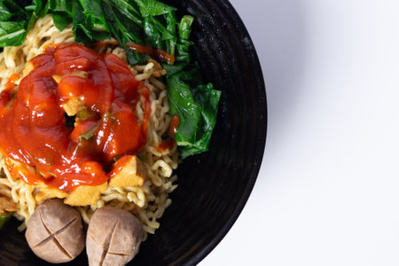 Top view of Indonesian chicken noodles, mie ayam, served with vegetables, meatballs, and spicy sauce in a black bowl on a white background.の写真素材