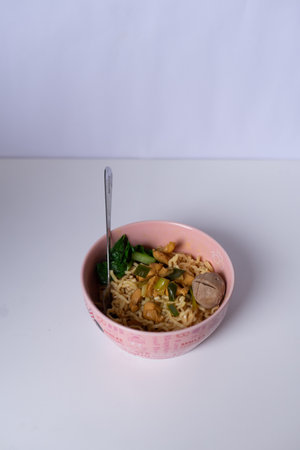 A tasty bowl of chicken noodles with greens and a meatball. Served in a pink bowl with a spoon, set against a clean white backdrop.の写真素材