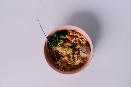 A bowl of chicken noodle topped with green vegetable and meatball on a white surface. Shot from aboveの写真素材