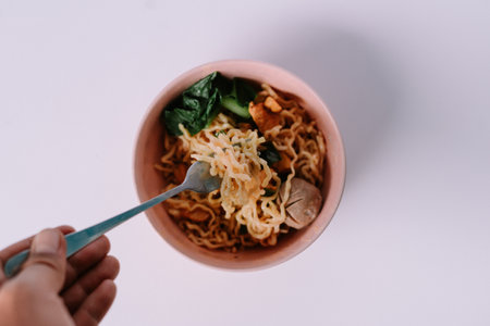 A savory noodle meal is held by a hand, ready to be eaten. The dish includes meatballs and green vegetables.の写真素材