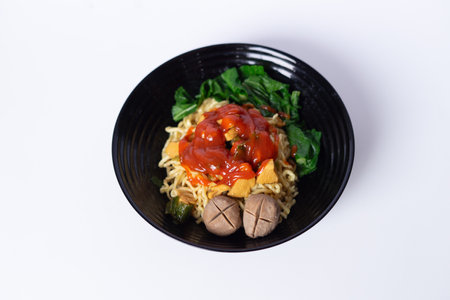Noodle dish with chili sauce and leafy greens in black bowl. Traditional flavor, top view on clear backdrop.の写真素材