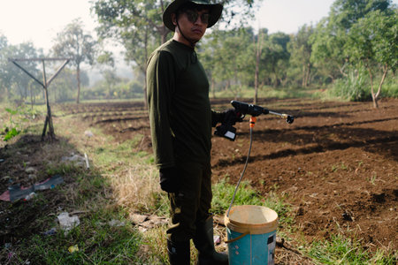 A farmer walking into the field carrying a sprayer, ready to irrigate or water the crops manually. Agricultural work in progress.の写真素材