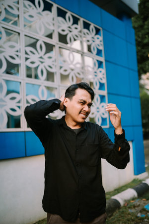 A young man is experiencing intense neck discomfort. He is standing by a building with a blue facade...