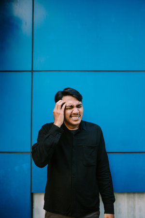 A young man is experiencing headache or migraine against a blue wall background. Showing stress and painの写真素材