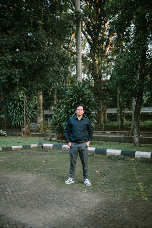 A content Asian man stands in an outdoor park setting, casually dressed and enjoying the serene environmentの写真素材