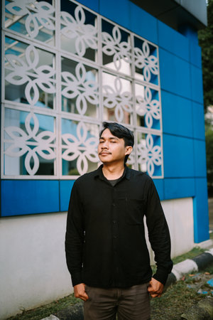 Portrait of a contemplative young man standing in front of a modern building. He exudes confidence and style in this urban scene. Daydreaming captured with sunlight.の写真素材