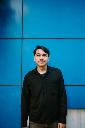 Pensive young man in casual clothing stands against a vibrant blue patterned wall looking awayの写真素材