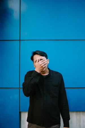 Upset man covering his face with hand against a blue wall. Feeling exhausted, stressed and...