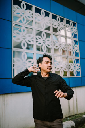 Contemplative asian man gestures. In background is building facade with white floral pattern on dark...