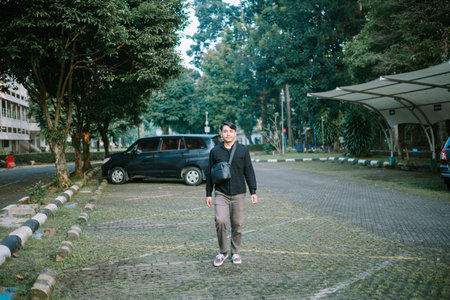 Young man casually walking outdoors in a parking area surrounded by trees. Relaxed and confident...