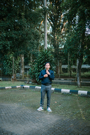 Asian man adjusting his shirt outdoors. Mature man in casual clothing amidst lush foliage in a...