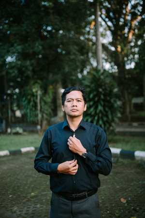 Portrait of a serious asian man buttoning his shirt outdoor. Male getting ready for a day at work, surrounded by natureの写真素材