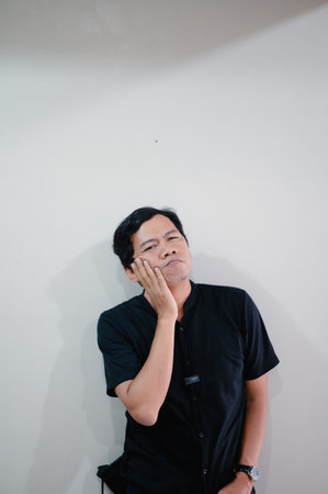 A man in a black shirt, deep in thought, places a hand on his cheek. The studio shot highlights his expression and contemplation against a clean white backdrop conveying a sense of pondering aの写真素材