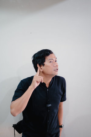 A portrait of a man in a black shirt, pointing at his ear as he concentrates, conveying a sense of listening, thought, and engagement, isolated against a plain white background.の写真素材