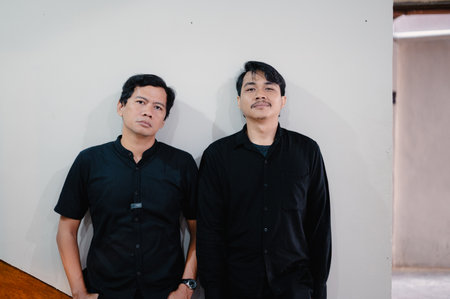 Two men are positioned closely together, leaning against a wall. They are dressed in black shirts, displaying a sense of unity and style. Their facial expressions are serious, suggesting focus orの写真素材