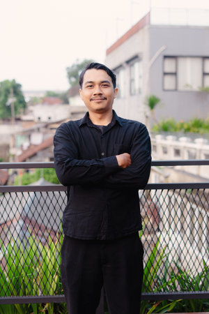 A smiling man in a black shirt and pants stands outdoors with arms crossed. He looks directly at the...