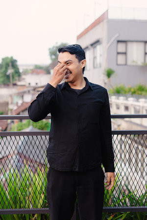 A portrait of a man smiling and touching his nose on a rooftop with an outdoor setting, he is wearing black shirt and pants.の写真素材