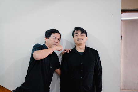 Two men in black shirts standing next to a white wall posing for a photo, one man looks assertive and touches the shoulder of the other smilingの写真素材