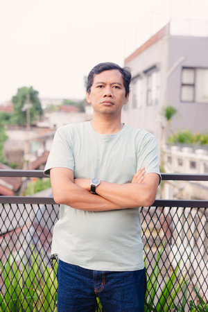 A middle-aged man stands on a balcony with his arms crossed, against an urban backdrop. His serious expression suggests contemplation and calm amidst the cityscape.の写真素材