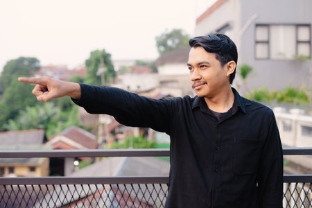 An asian man with a dark shirt is pointing with his right arm to the side, in an outdoor setting in a town and smiling with positive and cheerful expressionの写真素材