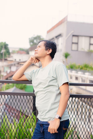 A man gazes upward thoughtfully while leaning on a railing, with a cityscape and overcast sky forming the backdrop of this contemplative moment.の写真素材
