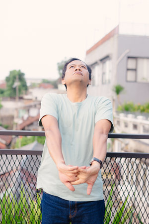 A man stands on a balcony, stretching and looking upwards. He appears relaxed, enjoying the outdoor ambiance. The setting is urban with a fence and a building in the background.の写真素材