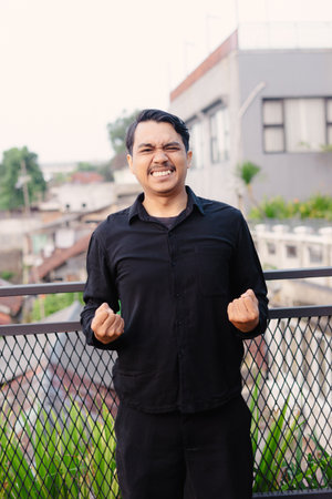 A man stands on a rooftop, fists clenched in a gesture of triumph. The background shows an urban landscape, suggesting the achievement is related to his environment. His expression shows a mixtureの写真素材