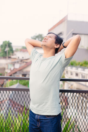 A man stands on a balcony, enjoying a moment of peace and contemplation. He gazes upwards, hands behind his head, embracing a sense of freedom and ease while observing the serene skylineの写真素材