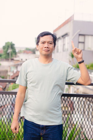 A middle-aged Asian man stands on a balcony, gesturing upwards with a raised finger, suggesting an idea or a point of interest. He's dressed casually, conveying a thoughtful expression.の写真素材