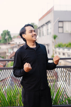 An elated man, bathed in the evening glow, joyfully celebrates on a rooftop balcony. With clenched fists and a radiant smile, he embodies triumph and urban delight, capturing the essence of success.の写真素材