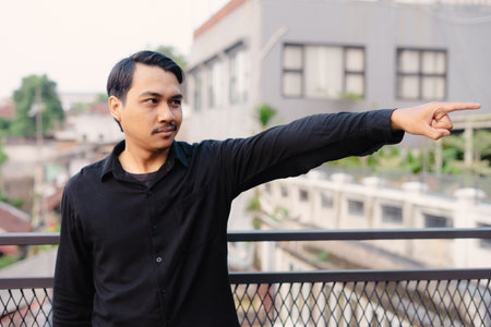 A man in a black shirt points in a direction, with a city background. The individual is outdoors, with a focus on his gesture and expression. The image captures a moment of interaction and directionの写真素材