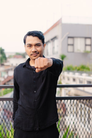 A man in a black shirt extends a clenched fist, conveying a sense of confidence, strength, and determination against a blurred urban backdrop with metal railing.の写真素材
