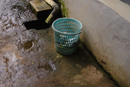 A close-up shot showcasing a turquoise plastic trash bin situated on a textured, weathered concrete surface, with a dark drainage opening nearby, suggesting an outdoor setting and a focus onの写真素材