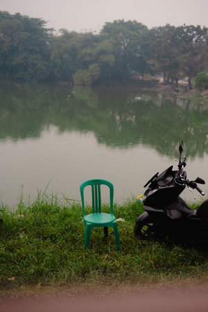 A vivid image capturing a serene lakeside setting. A green chair sits alongside a parked motorbike, with reflections in the tranquil water. Misty, natural surroundings enhance the tranquil atmosphere.の写真素材