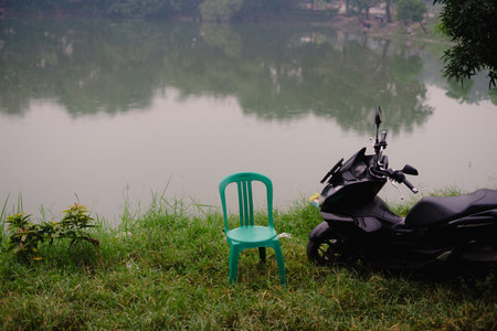 A tranquil scene of a lake with a motorcycle and chair on the grassy bank. The reflection in the water mirrors the trees and sky creating a peaceful atmosphere.の写真素材