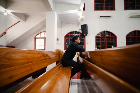 A man is seen sitting alone on a wooden bench in a church, lost in thought. The image captures a moment of introspection and spiritual reflection. The soft lighting and architectural details createの写真素材