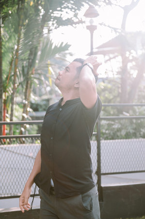 An Asian man in a black shirt looks up toward the sky while outside in a tropical garden setting....