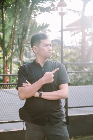 A stylish man stands outdoors, confidently pointing his finger while crossing his arms, suggesting direction or emphasis.の写真素材