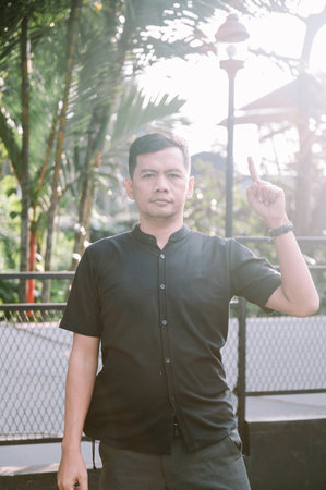 A man standing outside pointing his finger upwards. The background consists of lush greenery and a street light.の写真素材