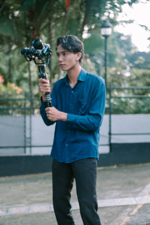 A young man holds and operates a camera and stabilizer outdoors. He is wearing a blue shirt and...