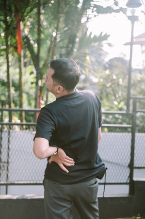 A man stands outside with a painful expression, holding his lower back. The image captures the discomfort of back pain.の写真素材