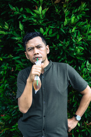 A man in a polo shirt is drinking iced tea from a plastic bag using a straw while standing in front of a green bush background.の写真素材