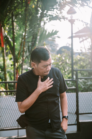 An adult man is experiencing chest pain while standing outdoors with trees and a bright sun in the background.の写真素材