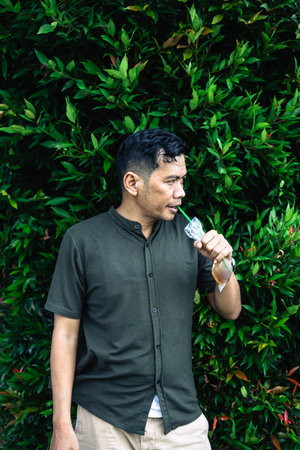 A man wearing a dark green shirt is seen outside, enjoying an iced drink with a green straw against a lush green foliage background.の写真素材
